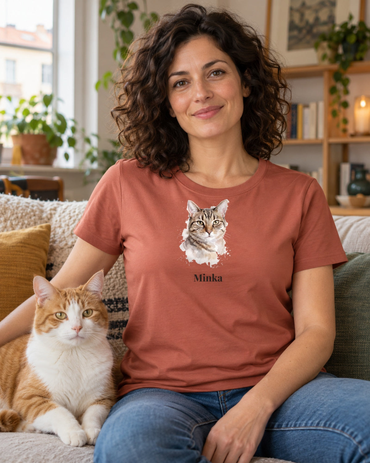 Frau im terrakottafarbenen T-Shirt mit Katzenportrait im Aquarell-Stil und Namen Minka, auf dem Sofa neben einer rot-weißen Katze