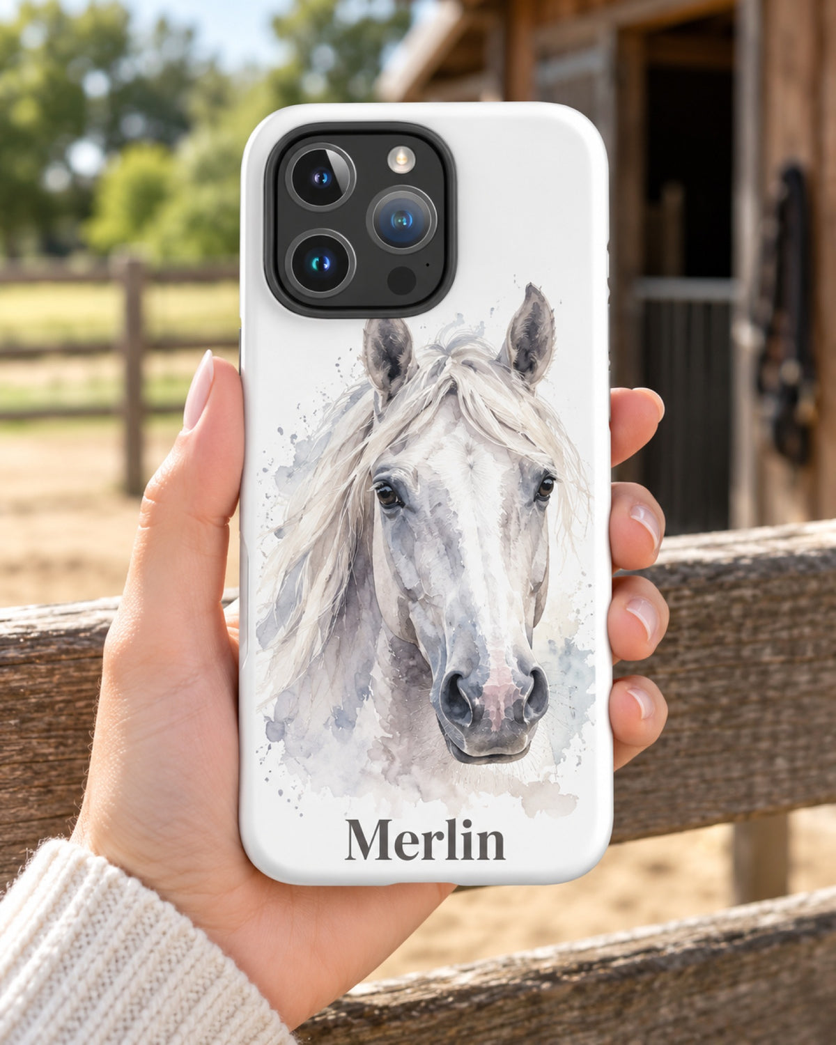 iPhone-Hülle mit Schimmel im Aquarell-Stil und Namen Merlin, eine Hand hält das Handy am Koppelzaun vor dem Stall