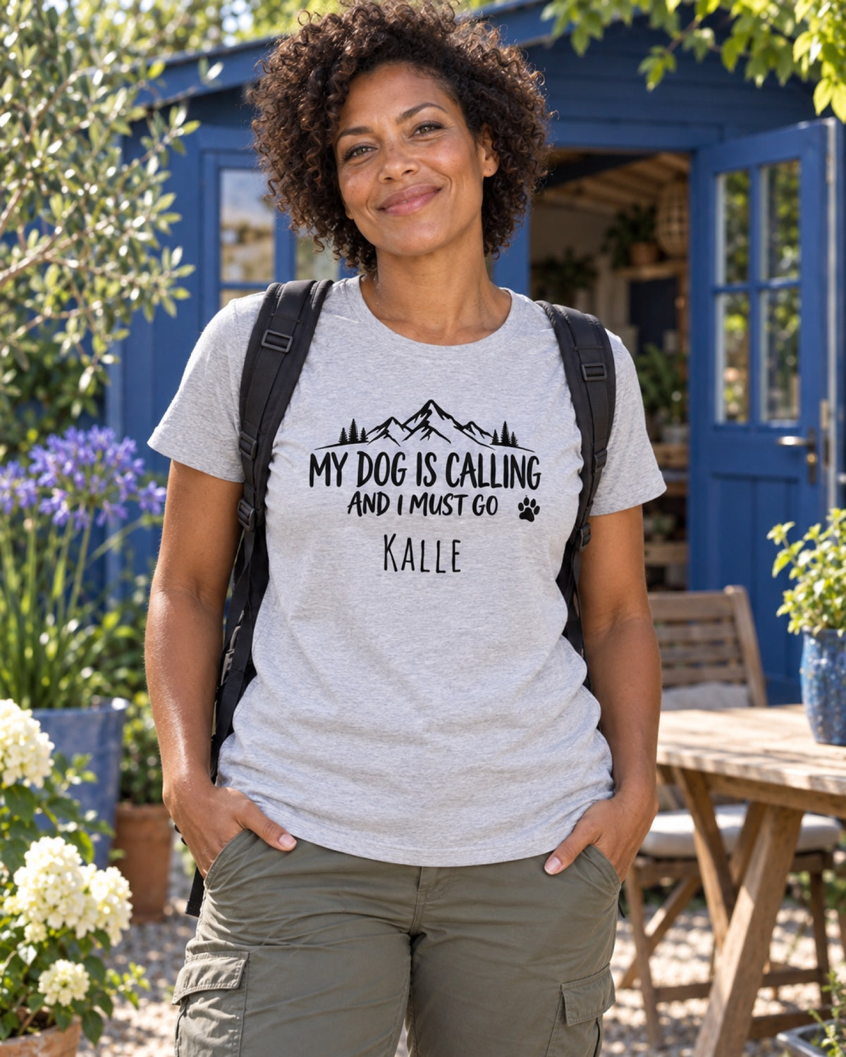 Frau im graumelierten My Dog Is Calling T-Shirt mit Beispielname Kalle vor einer blauen Gartenlaube