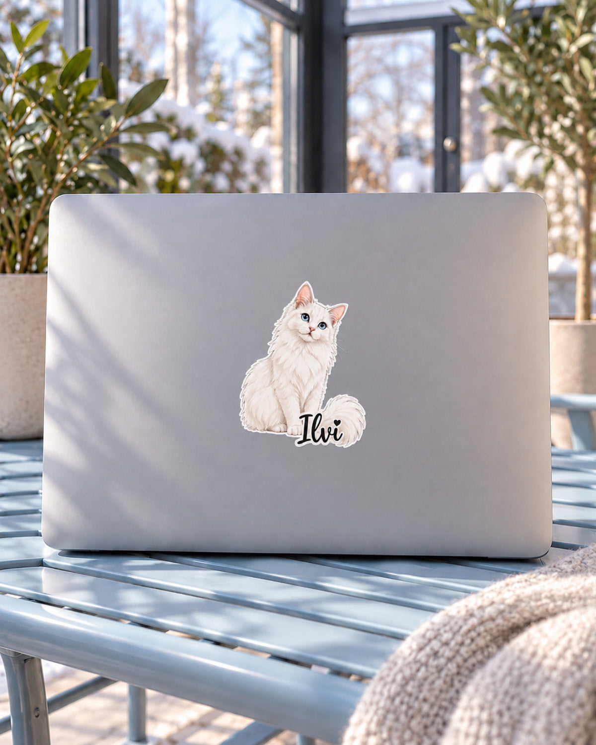 Vinylsticker 7,6 cm mit weißer Langhaarkatze im Original Stil und Namen Ilvi auf einem grauen Laptop auf einer hellblauen Metallbank im Wintergarten